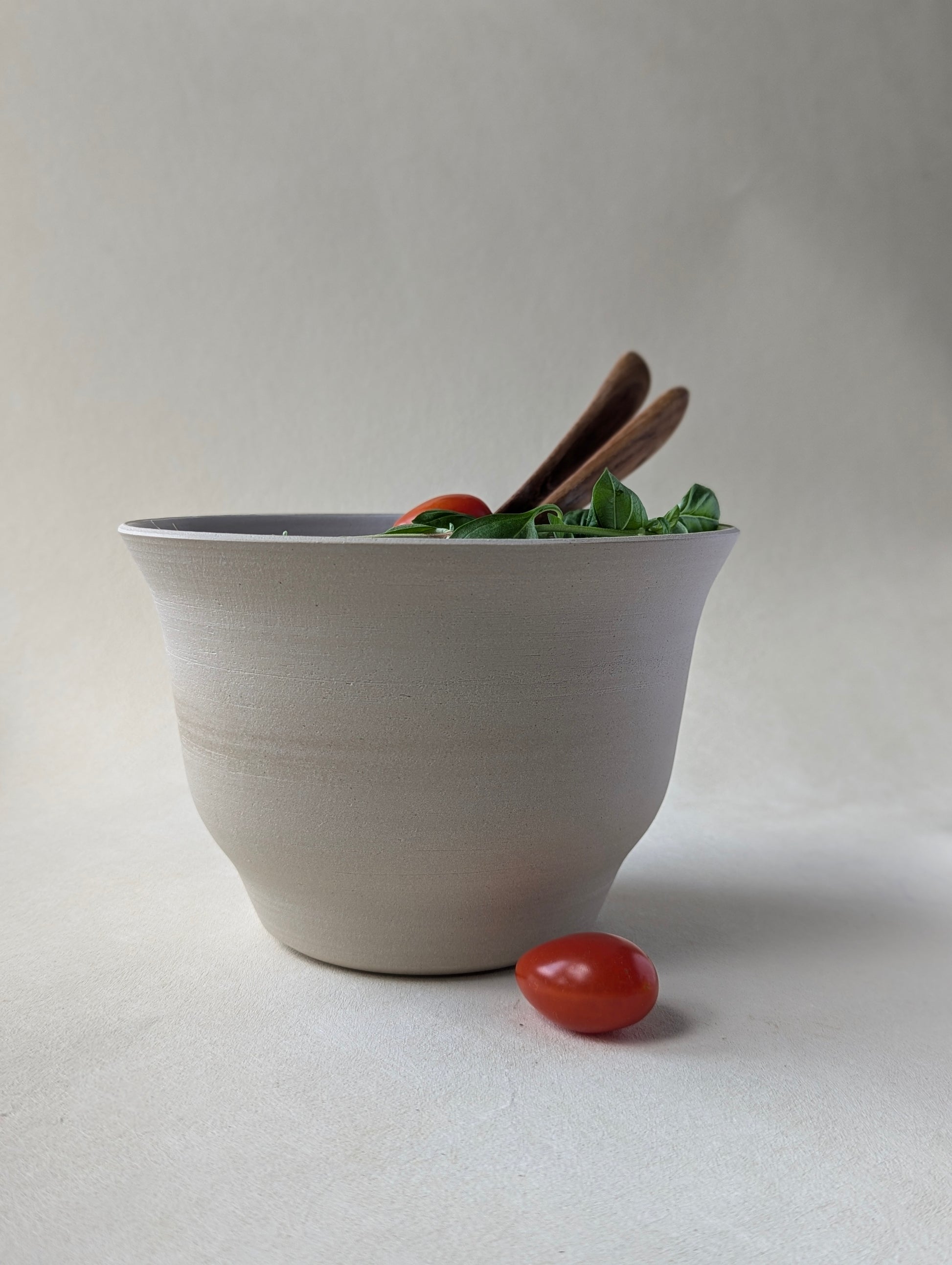 Handmade stoneware salad bowl with a glossy white interior and raw exterior, filled with fresh salad. Wooden salad spoon and fork are placed inside, showcasing the bowl’s elegant design and contrast finish.