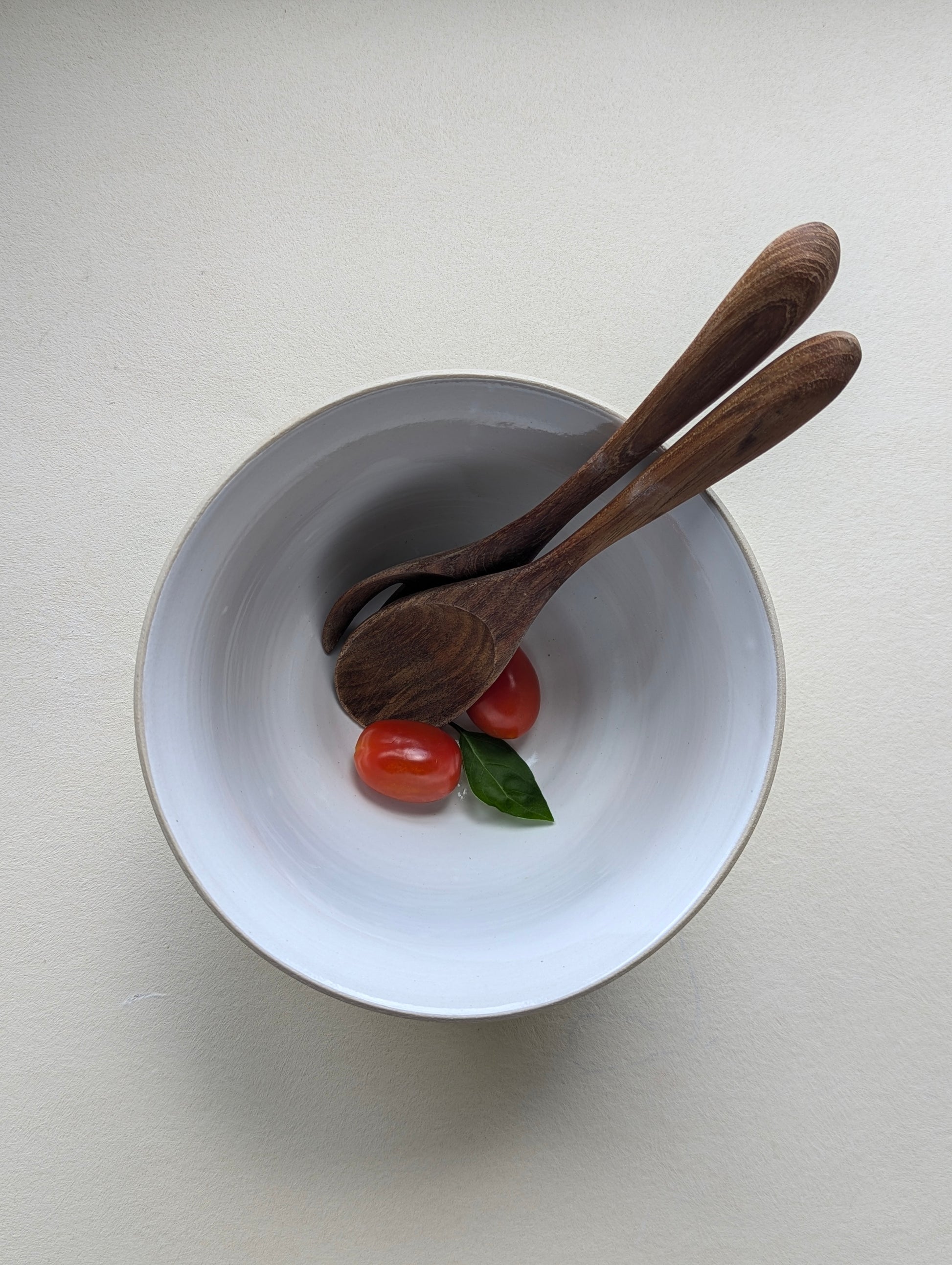 Top view of a handmade stoneware salad bowl with a glossy white interior, featuring a wooden spoon and fork inside, along with two cherry tomatoes for size reference.
